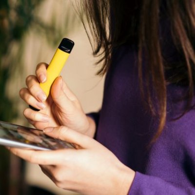 A young woman in profile with long dark hair falling across her face, holding a vape and a mobile phone.